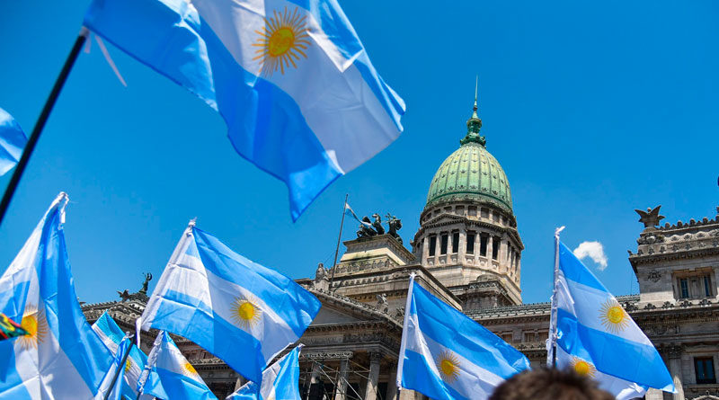 Banderas de Argentina siendo ondeadas por personas congregadas en la Plaza del Congreso de Argentina, ubicada en la Ciudad de Buenos Aires