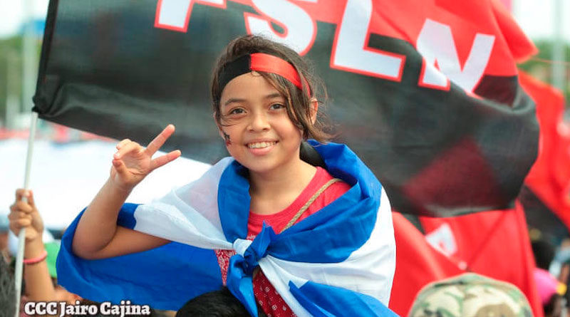 Jovencita saludando con la bandera de Nicaragua y el Frente Sandinista en su espalda